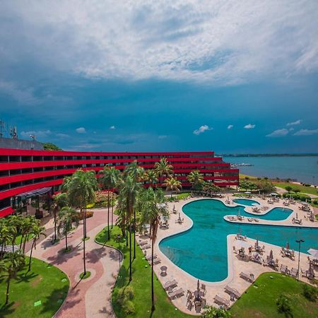 Hotel Royal Tulip Brasilia Alvorada Exterior foto The photo shows a modern hotel building with a striking red exterior, surrounded by lush greenery. In the foreground, there is a large, creatively shaped swimming pool, with lounge chairs arranged around it. The backdrop features a body of water, pos