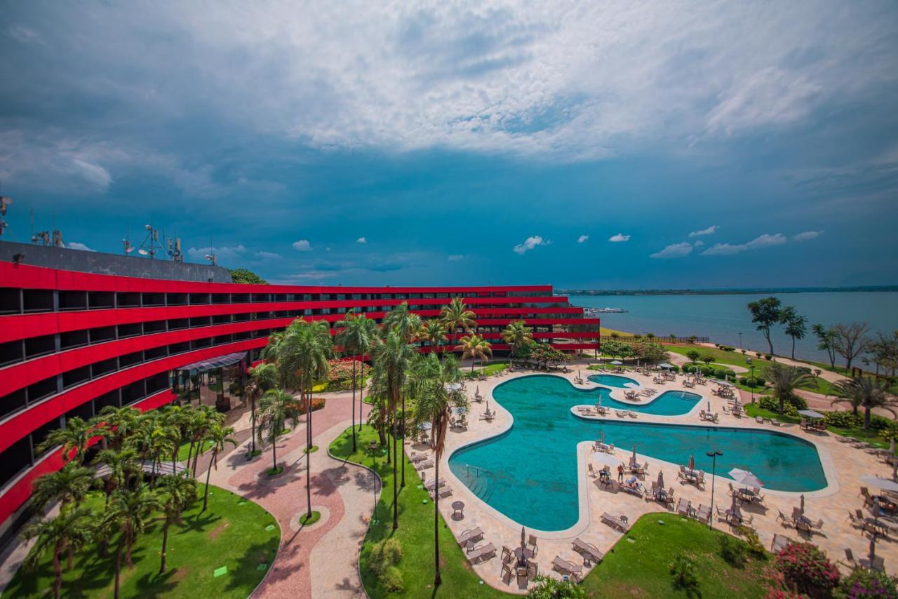 Hotel Royal Tulip Brasilia Alvorada Exterior foto The photo shows a modern hotel building with a striking red exterior, surrounded by lush greenery. In the foreground, there is a large, creatively shaped swimming pool, with lounge chairs arranged around it. The backdrop features a body of water, pos