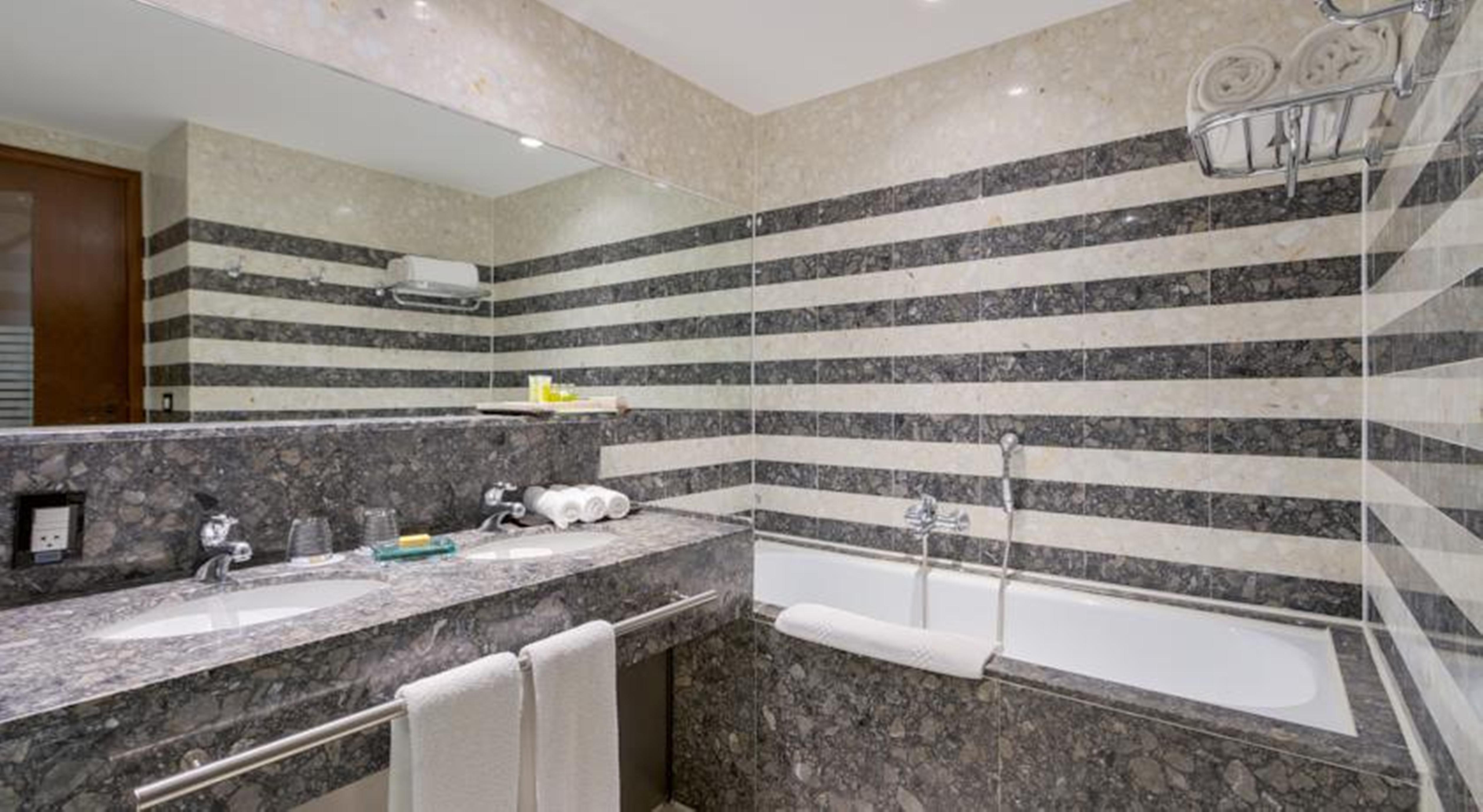Hotel Royal Tulip Brasilia Alvorada Exterior foto The photo shows a modern bathroom featuring a combination of marble and stone finishes. The walls are decorated with horizontal stripes in various shades of gray and cream. There is a bathtub with a white exterior, and a showerhead is mounted on the 