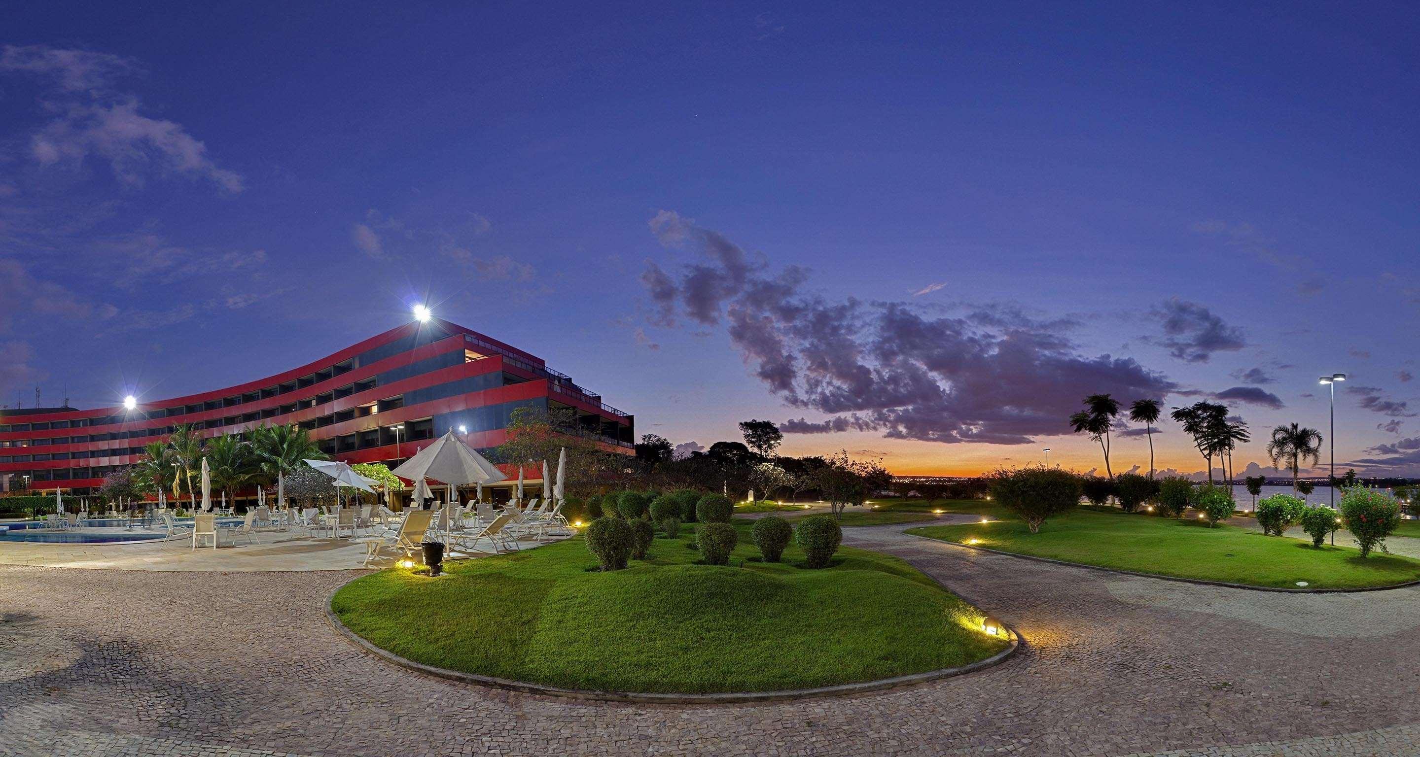 Hotel Royal Tulip Brasilia Alvorada Exterior foto The photo depicts a modern building, possibly a hotel or resort, with a striking red exterior. It is set in a landscaped area that features well-maintained greenery and ornamental plants. In the foreground, there are lounge areas with umbrellas, sugg