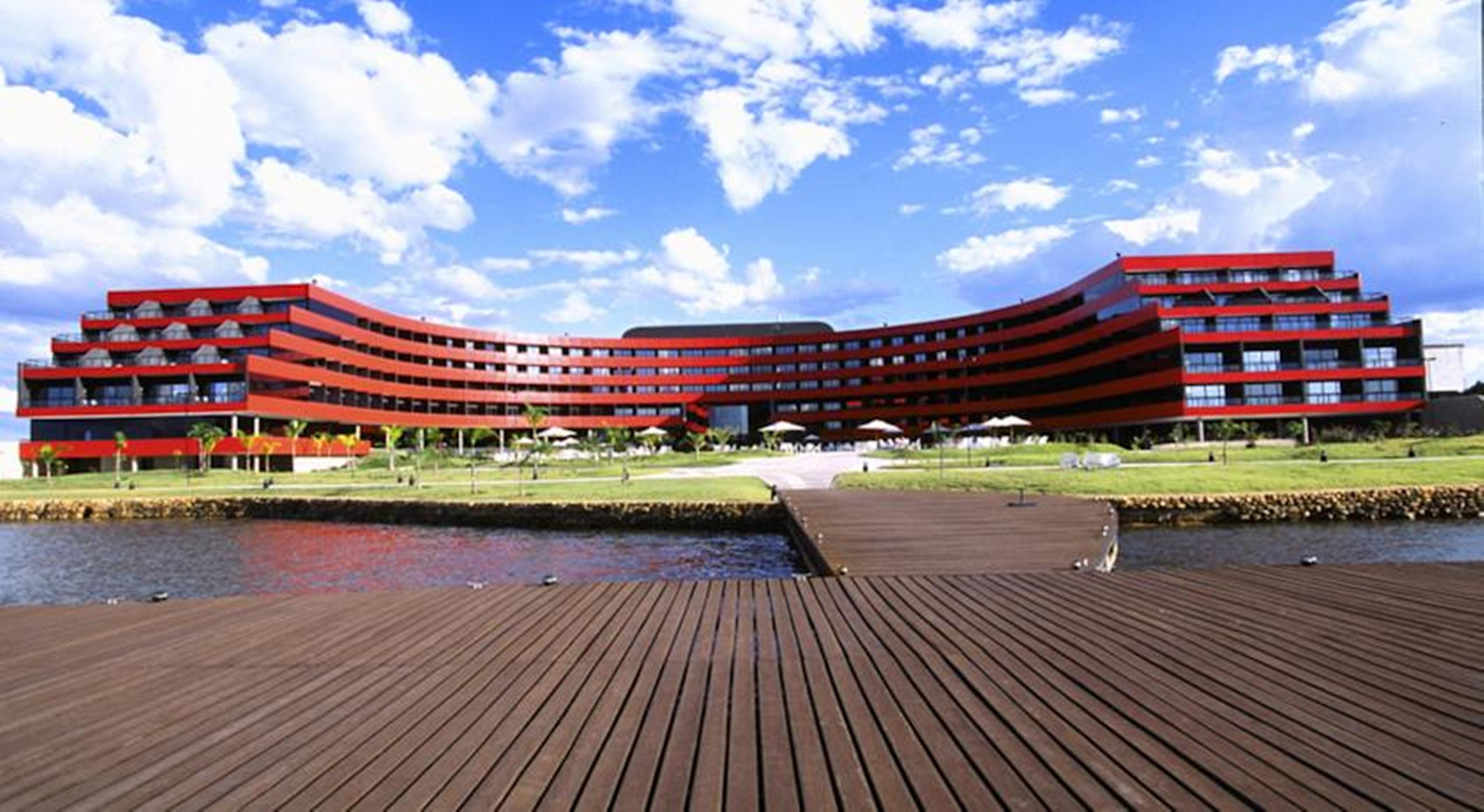 Hotel Royal Tulip Brasilia Alvorada Exterior foto The image shows a modern, curved building with a striking red facade, situated near a body of water. The architecture features multiple levels with large windows that create a sleek and contemporary appearance. In front of the building, there is a wo