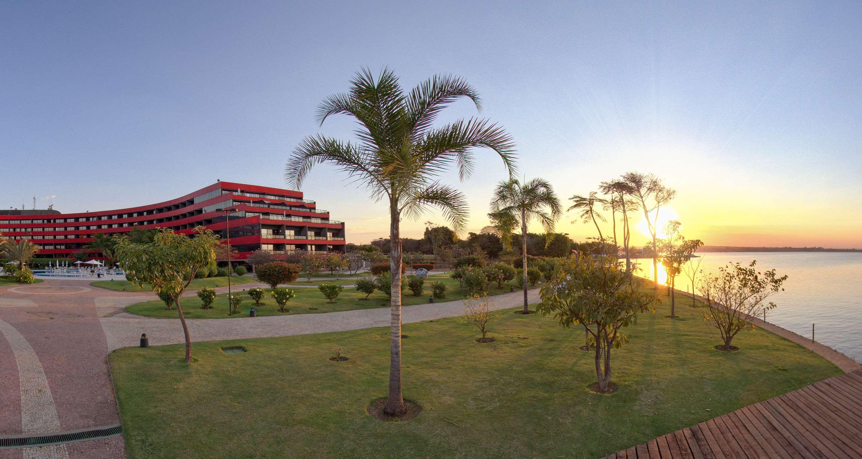 Hotel Royal Tulip Brasilia Alvorada Exterior foto The photo depicts a scenic view featuring a red multi-story building alongside a serene waterfront. In the foreground, there are several palm trees and lush green lawns, creating a tropical atmosphere. The sun is setting in the background, casting wa