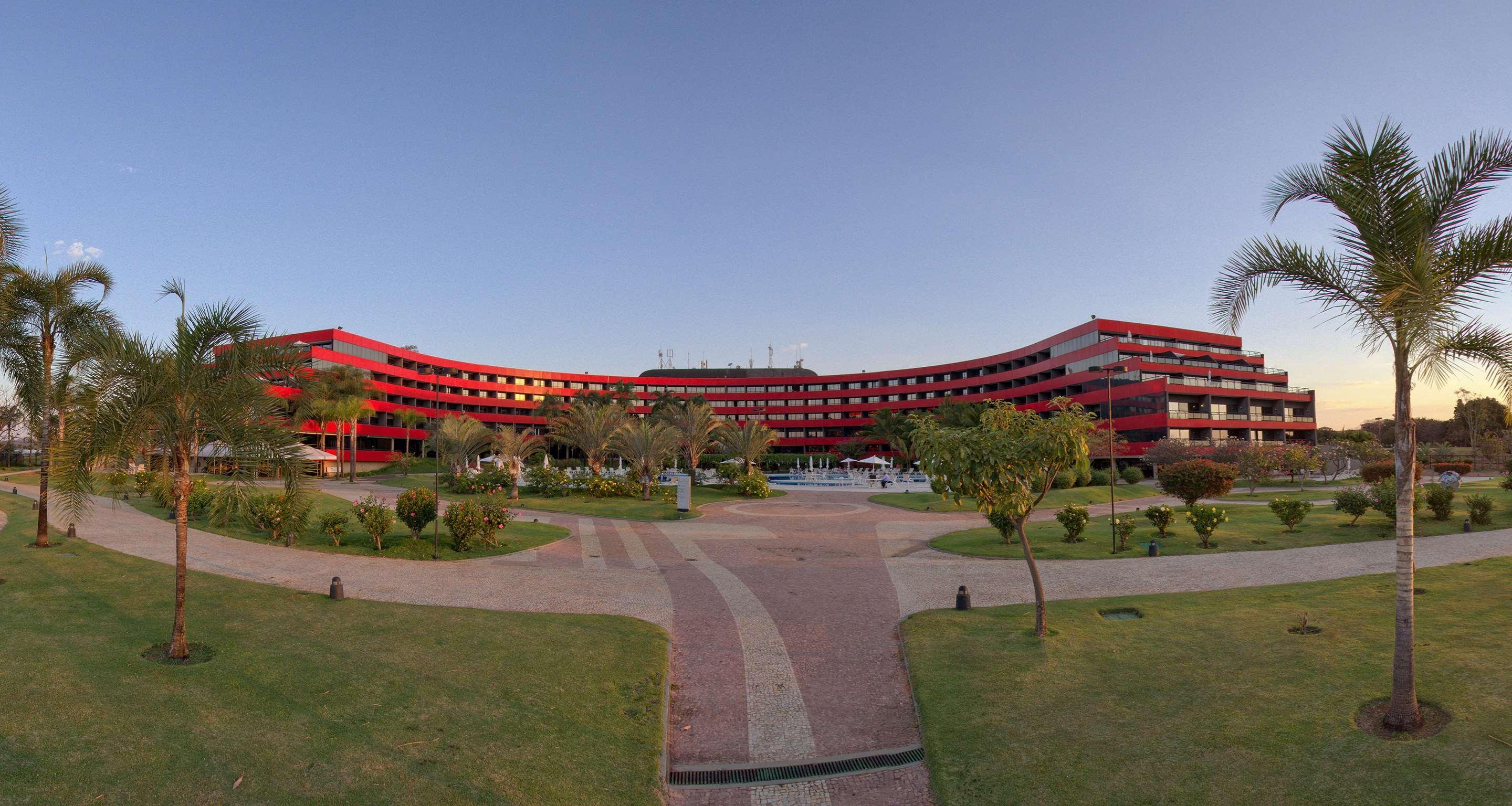 Hotel Royal Tulip Brasilia Alvorada Exterior foto The photo shows a large, curved building with a vibrant red exterior. It appears to be a hotel or resort, surrounded by well-maintained gardens and green spaces. The landscape includes manicured lawns, small trees, and pathways, suggesting a pleasant