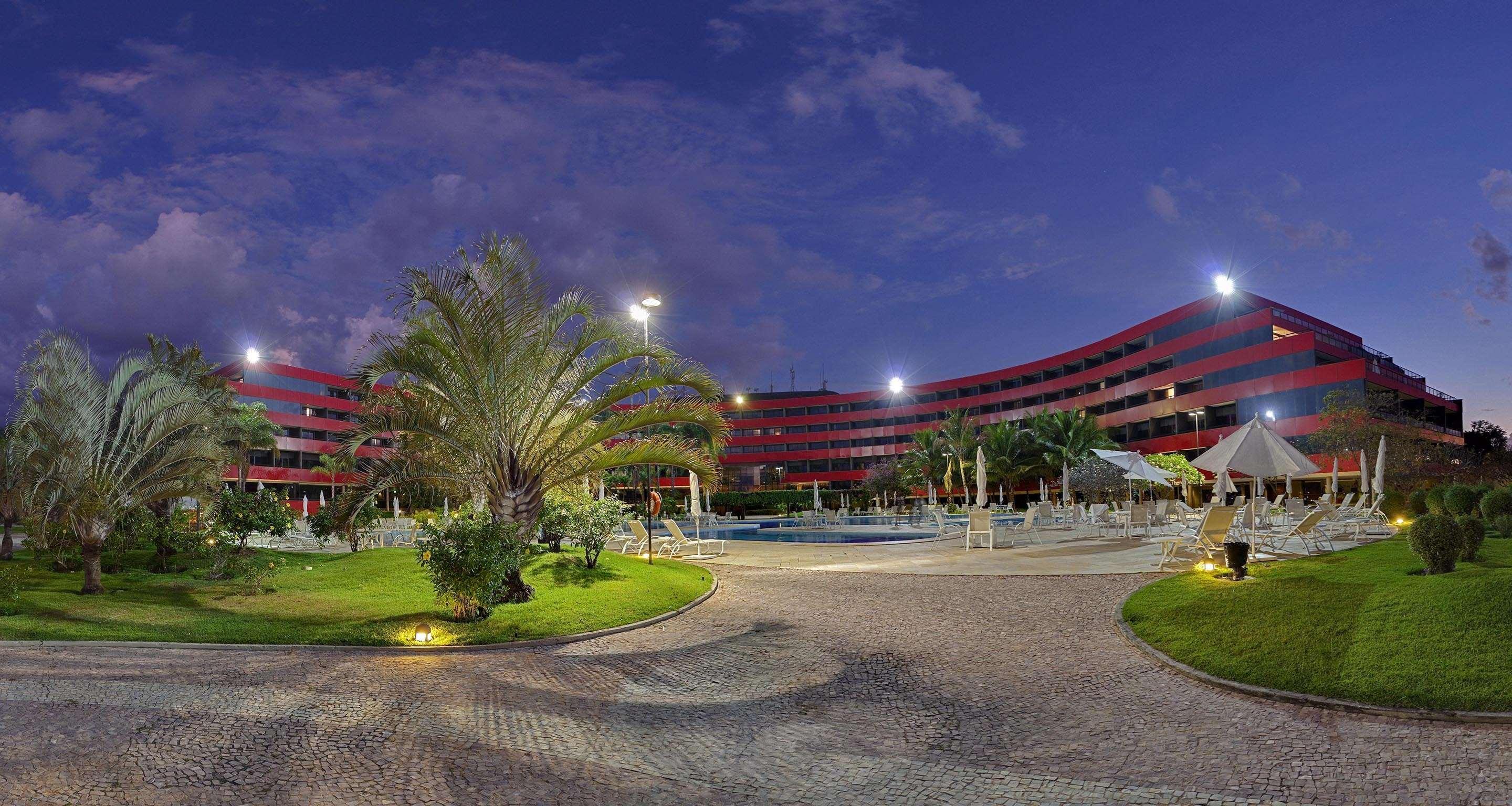 Hotel Royal Tulip Brasilia Alvorada Exterior foto The photo shows a panoramic view of a hotel or resort complex, characterized by its modern architectural design with smooth, curved lines and a vibrant red color. It’s set in a landscaped area featuring palm trees and green grass. The surroundings in
