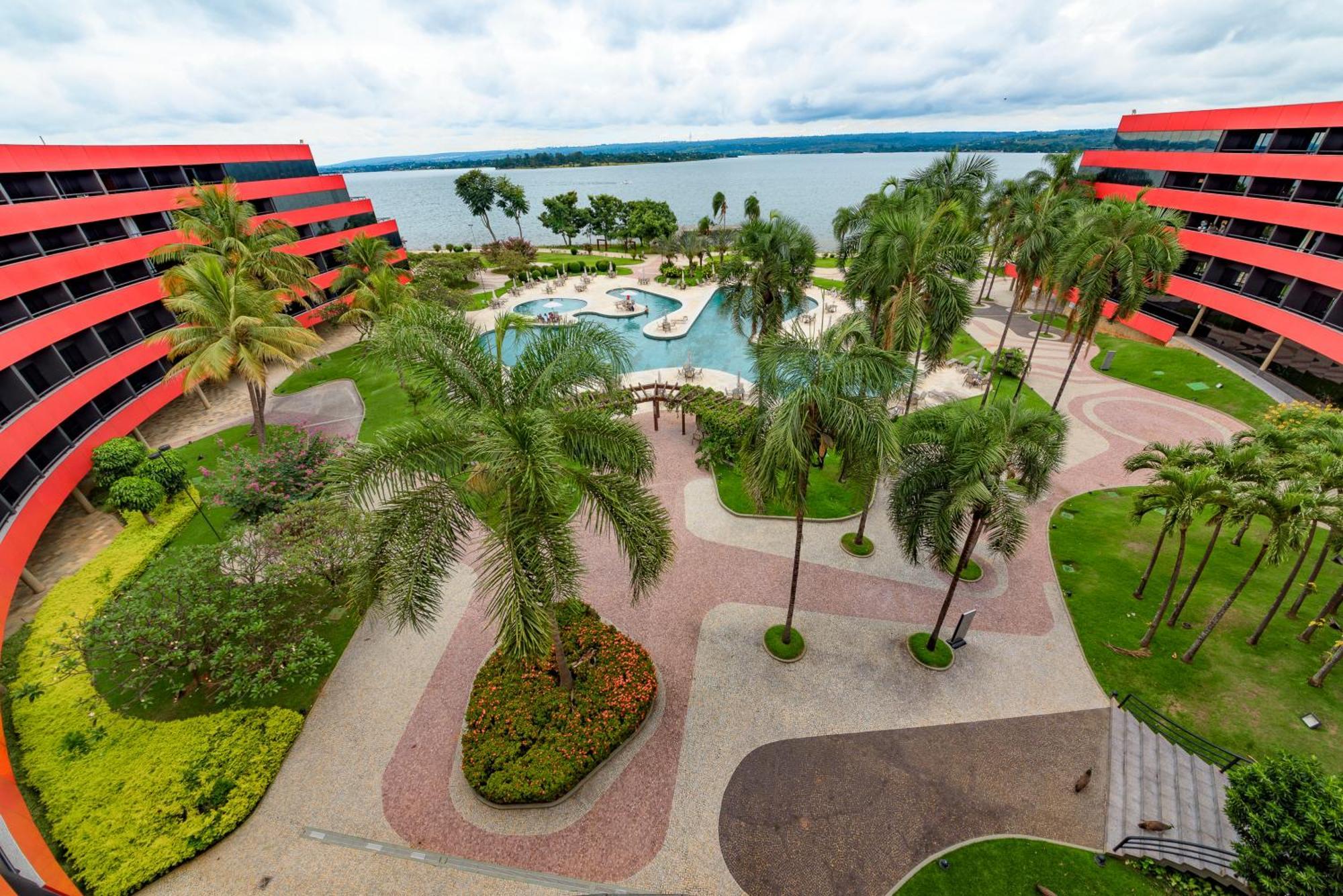 Hotel Royal Tulip Brasilia Alvorada Exterior foto The photo shows a scenic view of a courtyard area in a residential complex. There are multiple red buildings surrounding a pool area, which is surrounded by palm trees and landscaped gardens. The pool features a curvy design, and there are pathways m